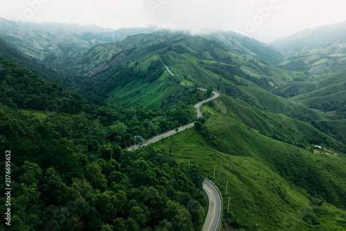 Aerial view of a winding asphalt road cutting through lush green mountains and tropical rainforest.