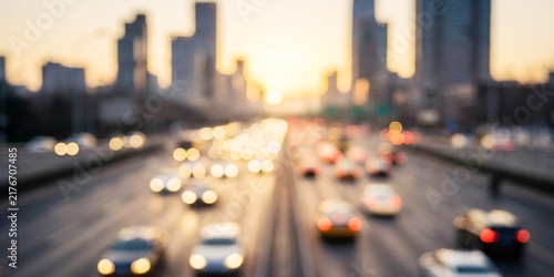 A blurred view of a busy highway filled with cars, set against a backdrop of tall city buildings during sunset, creating a vibrant urban atmosphere.