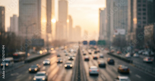 A blurred cityscape during sunset, featuring a busy road with vehicles. Tall buildings align the street, creating a vibrant urban atmosphere.