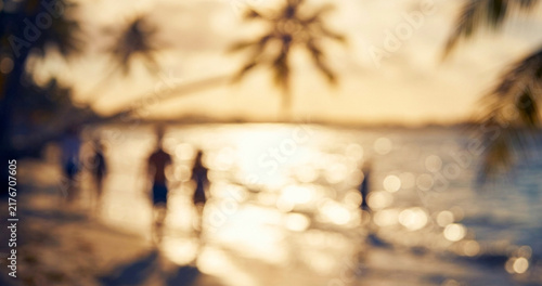 A serene beach scene at sunset, featuring blurred silhouettes of people walking along the shore surrounded by palm trees.