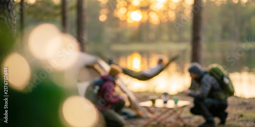 A serene camping scene by a lake at sunset, with a group of friends engaged in various activities, surrounded by nature and trees.