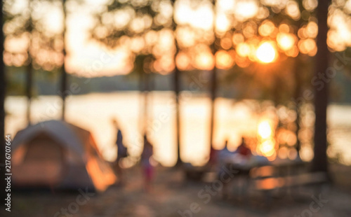 A blurred sunset scene at a lakeside campsite, featuring silhouettes of people, a tent, and trees, creating a tranquil outdoor atmosphere.