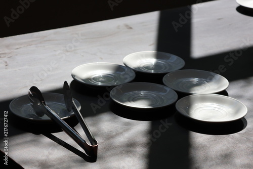 Sunlight casts soft shadows across minimalist tableware. Five clear plates rest beside metal tongs. Neutral tones evoke calm, quiet dining moments. Ideal for design, lifestyle, or editorial projects