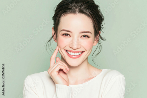 Portrait of beautiful young woman pointing finger to cheek smiling cheerfully with white teeth against green background for beauty skincare cosmetics and wellness concept