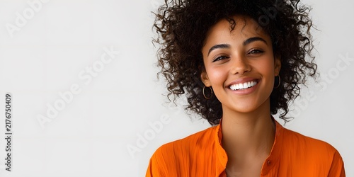 Cheerful young woman with curly hair smiling in orange shirt against light background