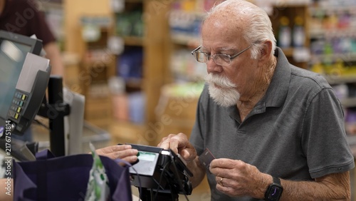 Картината върху платно An elderly man is at the grocery store using his card to pay for his items at the checkout