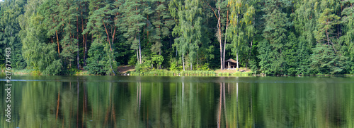 Fotografie Coniferous Forest Reflecting in Calm Lake Surface with Wooden Shelter on Sunny S
