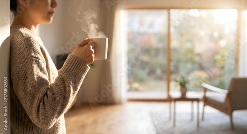 Woman in cozy wool sweater holding steaming hot mug of tea by living room window on cold winter morning