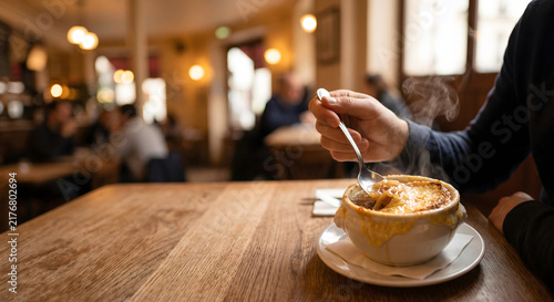 Close up of hand dipping spoon into steaming French onion soup topped with melted cheese in cozy bistro