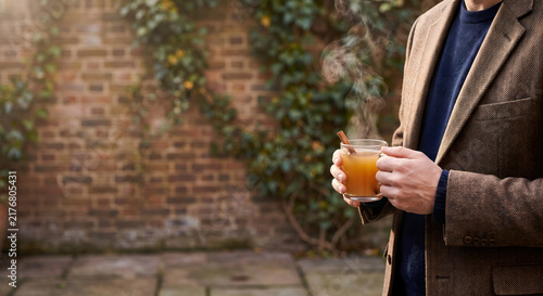 Person holding a glass mug of hot spiced cider with cinnamon in a courtyard