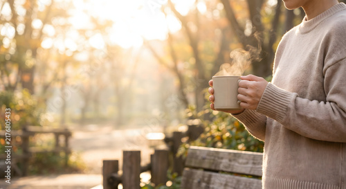 Person holding a steaming ceramic mug outdoors in a sunlit park in autumn