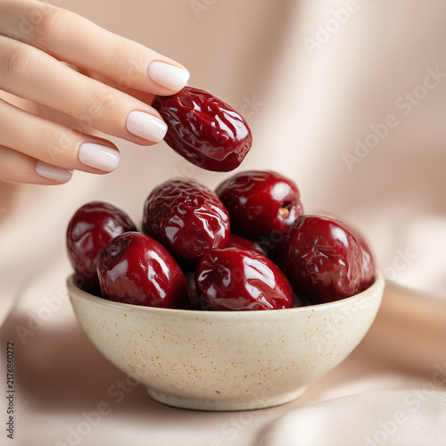 Close-up of a manicured hand selecting a glossy red date from a ceramic bowl filled with ripe dates, styled with soft beige tones and elegant natural light.