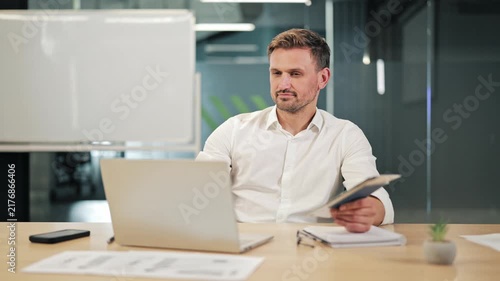 Dedicated business person diligently works at office desk. Uses laptop and reviews documents, focus and corporate responsibility. Professional environment supports concentration and productivity.
