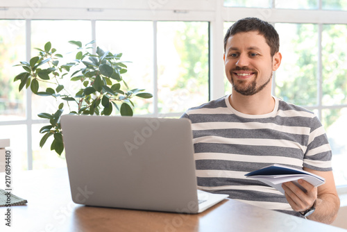 Young male teacher with laptop and notebook giving online lesson at home