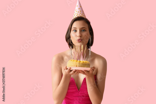 Beautiful woman in party hat with Birthday cake making wish on pink background
