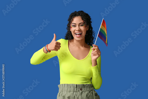 Happy African-American woman with rainbow flag showing thumb-up on blue backg...