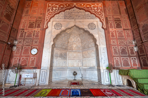 Intricate white marble mihrab prayer niche with Arabic calligraphy framed by red sandstone wall inside prayer hall at Jama Masjid, Delhi, India.