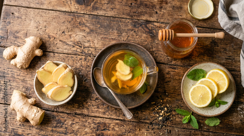 Top-down view of a glass cup of hot ginger tea with mint leaves, lemon slices, honey jar, and whole ginger roots on a rustic wooden table.