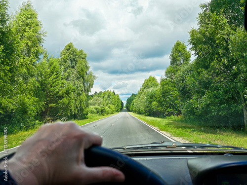 Driver POV from inside a car shows sunlit two-lane highway runs straight through dense summer forest beneath a clear blue sky with thin wispy clouds.