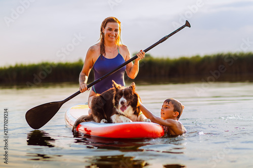 Young boy swimming in the river and petting his dog who sits on a paddleboard held by his smiling mother during a sunset.