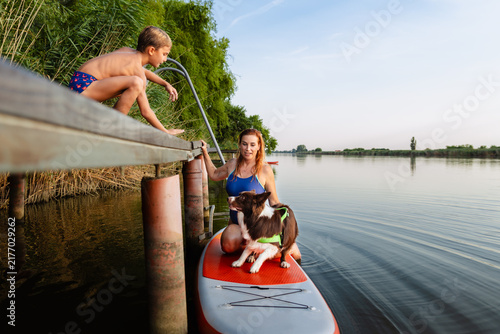 Smiling mother kneeling on paddleboard while son calls dog from wooden dock by the river.