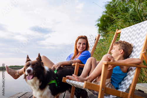 Smiling mother sitting in wooden chair petting her dog next to son on river dock.