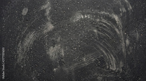 Top-down view of a dark, textured asphalt surface with white chalky streaks and dusty marks across the grainy pavement.