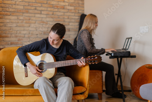 Teen girls making music indoors at home, one focused on guitar and the other on keyboard. Cozy living room with brick wall and warm furniture. Creativity, bonding, teamwork and leisure concepts. 
