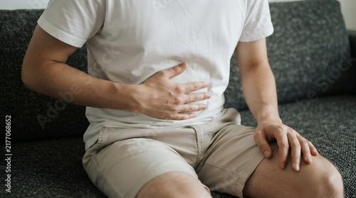 Stomach ache and abdominal pain, man holding belly with digestive issues, male patient suffering from cramps on sofa
