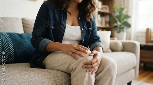 Woman holding painful knee on sofa, female suffering from joint pain at home, concept of arthritis and physical injury