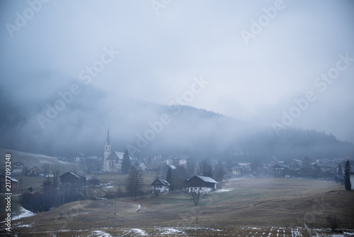 Wide shot of foggy alpine village with tall church spire surrounded by mist covered hills and light snow in winter. Atmospheric winter landscape in Gosau, Austria.