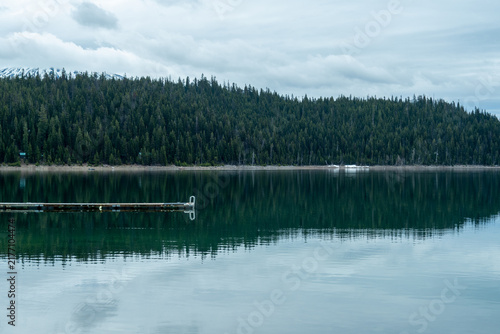 Canvas Print Serene Elk Lake scene with a floating dock and mirror-like water reflecting dense pine forest and distant snow-capped peaks beneath overcast skies