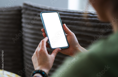 Woman Sits On The Couch At Home, Holding A Smartphone With A Blank White Screen In Hands