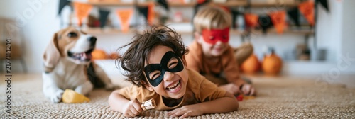 Two children and a dog wearing halloween masks laughing together on a carpet with pumpkins and decorations at home for seasonal background