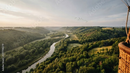 Hot air balloon basket overlooking winding river valley with forest at sunrise with balloon floats with floats above green