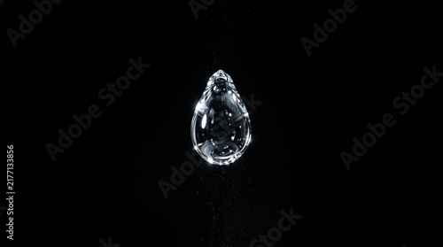 Close up of a single clear water drop falling against a stark black background with tiny particles