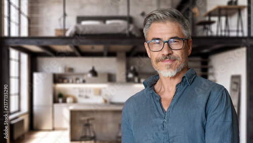 Portrait of confident senior man in shirt standing in modern loft apartment, smiling at camera. Gray haired mature guy with glasses and beard, casual style. Industrial interior, kitchen behind.