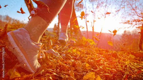 LENS FLARE, DOF, LOW ANGLE, CLOSE UP: Unrecognizable female runner moves through fallen autumn leaves while warm sunlight shines across the forest floor. Jogging amid flying dry leaves in fall season.