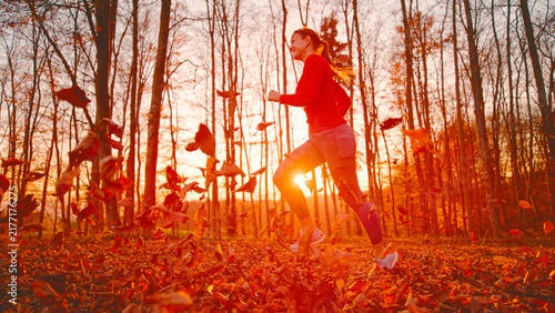 LENS FLARE: Cheerful young woman jogs through a colourful autumn forest while dry leaves swirl around her and warm low sunlight shines through bare trees. Outdoor exercise, movement and seasonal joy.