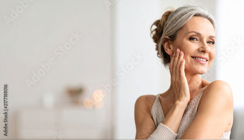 Confident middle aged woman touching her face with gentle smile and natural makeup indoors