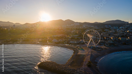 aerial drone shot of a Sunrise over Marseille’s waterfront with Ferris wheel, calm shoreline, rocky jetty, and golden mountains creating a serene coastal cityscape