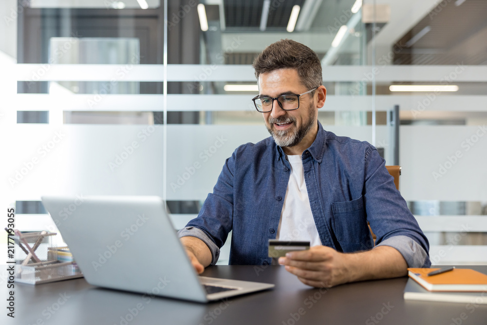 Obraz premium Man holding a credit card while using a laptop for secure online shopping and e-commerce transactions from a modern office desk, confident and smiling during remote work
