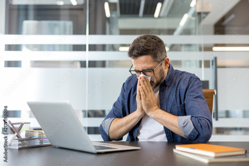 Obraz premium Businessman feeling unwell with cold or flu symptoms, blowing nose into a tissue while sitting at his office desk with a laptop, managing illness in a workplace environment