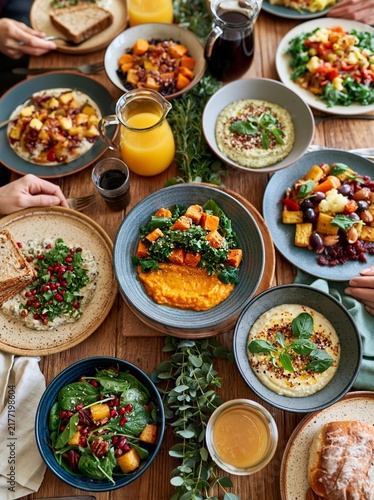 Group of people gathered around a wooden table, enjoying a meal together. there are multiple plates and bowls filled with different types of food, including salads, sandwiches, and dips.