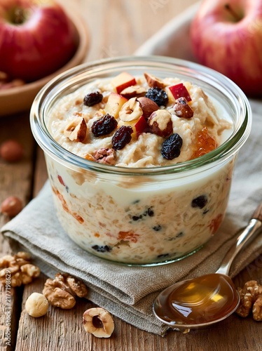 Glass jar filled with a creamy oatmeal mixture. the oatmeal is topped with sliced apples, raisins, and walnuts. the jar is sitting on a beige cloth napkin with a spoon resting on it.
