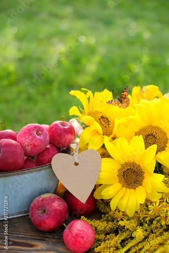 beautiful still life with red apples, paper heart shape blank and sunflowers close up on table in garden, natural background. farm harvest, summer end, autumn. fresh delicious healthy seasonal fruits