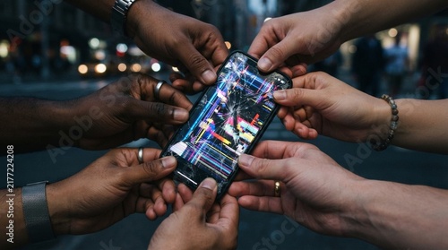 Multiple hands holding a cracked smartphone displaying digital glitch art