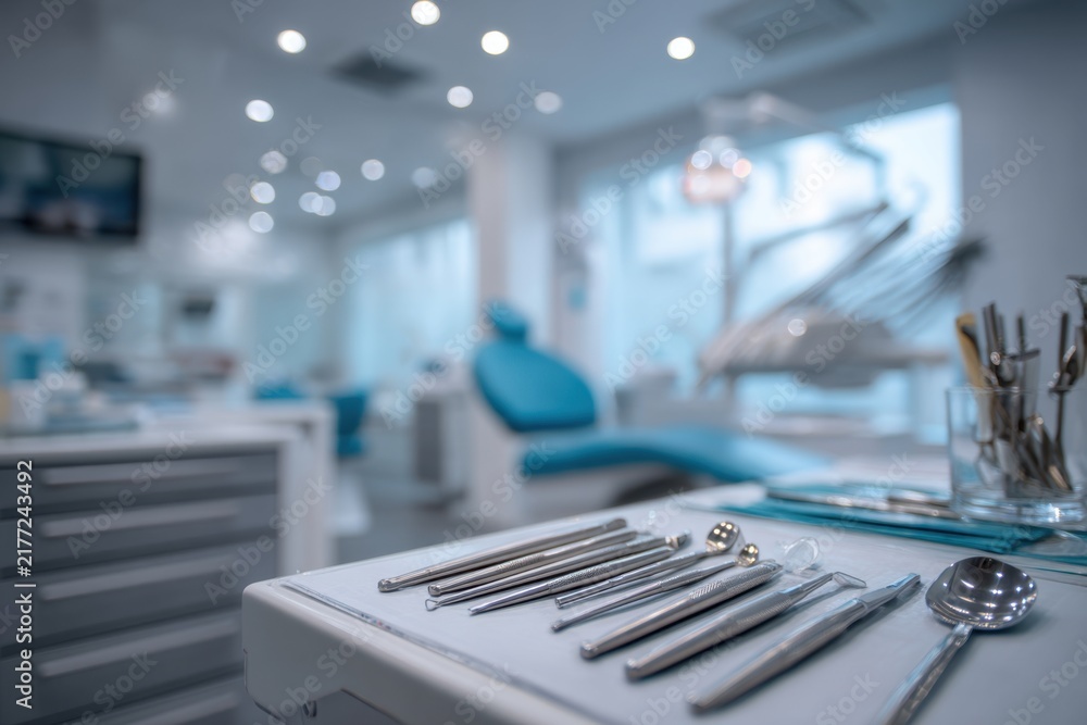Obraz premium Dental clinic interior featuring a tray of dental instruments in the foreground with a dental chair and equipment visible in the background