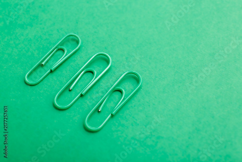 Monochromatic photo of three green paperclips arranged in a horizontal line on a green surface. Minimalist composition highlights repetition, geometry, and stationery.