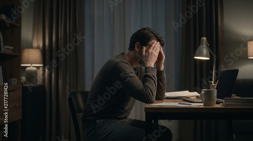 Horizontal photo of a tired man sitting at a desk in a dark room holding his head with both hands due to overwork